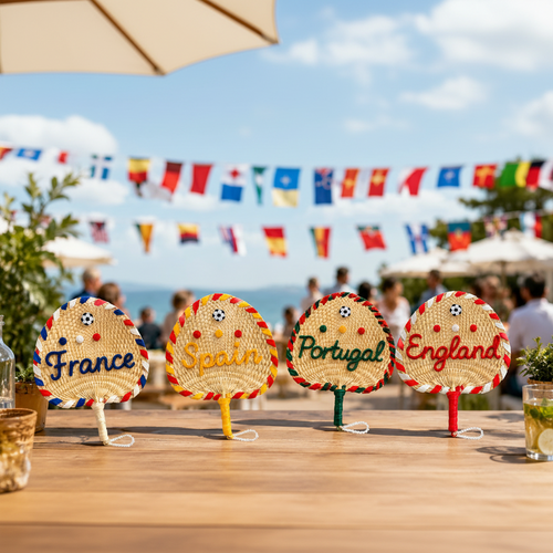 Four decorative fans labeled France, Spain, Portugal, and England on a table with a beach and flags in the background.